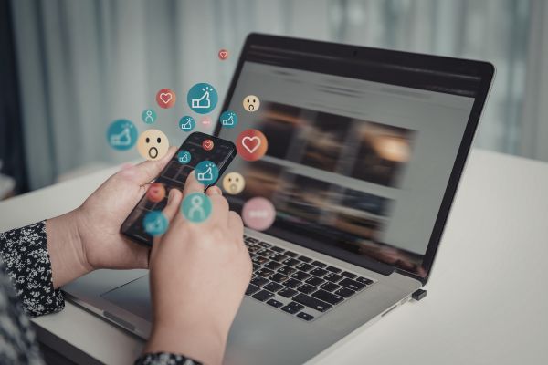 Person typing on a laptop with colorful social media icons and reaction emojis appearing from the keyboard