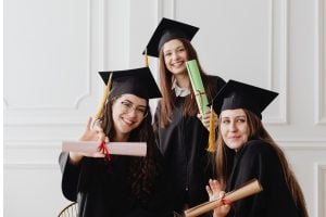 Three graduates in caps and gowns pose with rolled diplomas, smiling at the camera (pink, green, and brown diploma rolls).