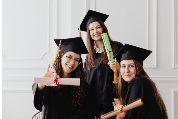 Three graduates in caps and gowns pose with rolled diplomas, smiling at the camera (pink, green, and brown diploma rolls).