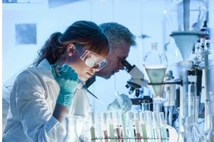 Two scientists in lab coats and safety goggles pipette liquids into a row of test tubes in a bright laboratory.