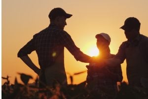 Three farmers in a field at sunset shake hands, signaling teamwork and partnership.