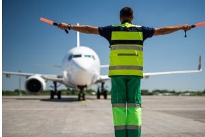 Ground marshal with neon safety vest and reflective pants, arms outstretched holding orange batons, directing a white airplane on the runway.