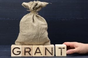 Burlap sack sits atop wooden blocks that spell GRANT, with a hand reaching in from the right.