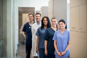 Five healthcare professionals—doctors and nurses—stand in a clinic hallway, smiling at the camera by patient rooms on the right.