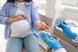 Pregnant woman seated with hand on belly as a clinician in blue gloves administers a vaccine in her upper arm.