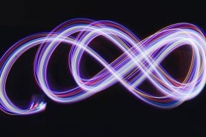 Long-exposure light painting forming a neon infinity symbol in purple, blue, and pink against a dark background.