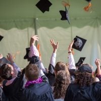 Graduates in caps and gowns throw their mortarboards in the air, shown from behind at a commencement ceremony under a tent.