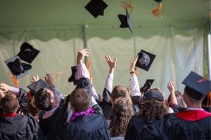 Graduates in caps and gowns throw their mortarboards in the air, shown from behind at a commencement ceremony under a tent.
