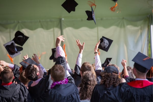Graduates in caps and gowns throw their mortarboards in the air, shown from behind at a commencement ceremony under a tent.
