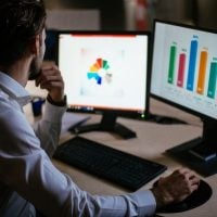 Man in a dress shirt sits at a desk, analyzing data on two monitors: left shows a colorful pie chart, right shows a bar chart.
