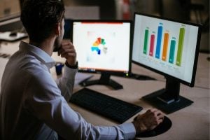 Man in a dress shirt sits at a desk, analyzing data on two monitors: left shows a colorful pie chart, right shows a bar chart.