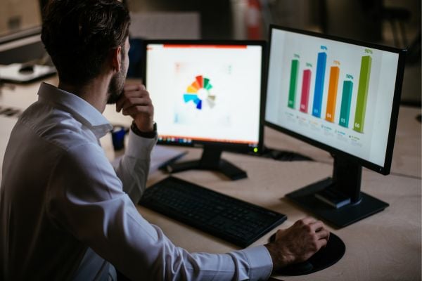Man in a dress shirt sits at a desk, analyzing data on two monitors: left shows a colorful pie chart, right shows a bar chart.