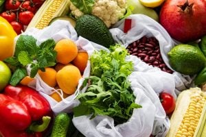 Assorted fresh produce in reusable cloth bags: leafy greens, peppers, tomatoes, corn, citrus, avocado and beans in a colorful display Fresh fruits and vegetables arranged in white bags: oranges, tomatoes, lettuce, corn, kidney beans and herbs Vibrant assortment of grocery produce in reusable bags: greens, corn, tomatoes, peppers, apricots and beans Colorful display of market produce in fabric bags: leafy greens, corn on the cob, peppers, citrus and legumes Bagged groceries with a variety of fruits and vegetables—greens, corn, tomatoes, beans, avocados and citrus—on a bright table