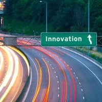 Road Transport and Innovation - fundsforNGOs Green highway sign reading 'Innovation' with an upward arrow over a curved multi-lane highway at dusk, showing light trails from passing cars toward the direction of innovation.