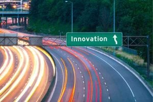 Green highway sign reading 'Innovation' with an upward arrow over a curved multi-lane highway at dusk, showing light trails from passing cars toward the direction of innovation.