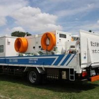 Large white utility truck with orange circular winches mounted on top, blue stripe and Japanese text, parked on a grassy field.