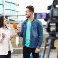 Young Journalist - fundsforNGOs Man in a denim jacket interviews a smiling woman with an orange microphone outdoors, camera recording them near a modern glass building.