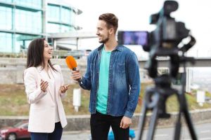 Man in a denim jacket interviews a smiling woman with an orange microphone outdoors, camera recording them near a modern glass building.