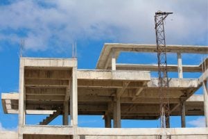 Concrete building frame under construction with exposed slabs and vertical columns; a rust-colored tower crane stands nearby against a blue sky.