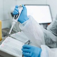 bioscience - fundsforNGOs Lab scientist in a white protective suit and blue gloves pipetting liquid into a 96-well microplate in a modern lab, with a yellow waste container in the background.