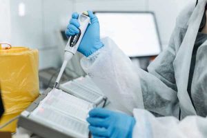 Lab scientist in a white protective suit and blue gloves pipetting liquid into a 96-well microplate in a modern lab, with a yellow waste container in the background.