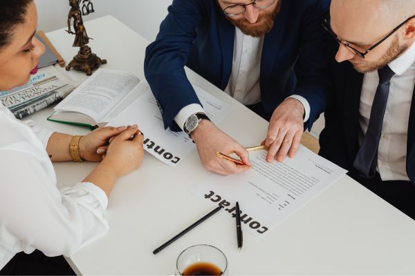 business 5 - fundsforNGOs Three professionals sign a contract at a white table, with documents labeled 'Contract' and pens nearby.