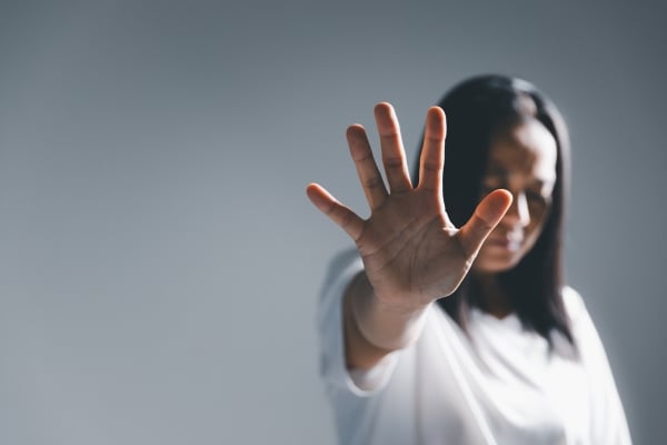 Woman in a white shirt raises an open hand toward the camera as a stop gesture, with her blurred face in the background.