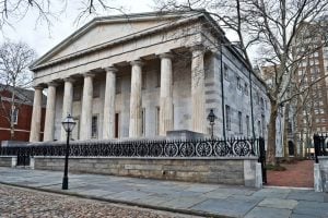 Neoclassical temple-style building with a row of tall stone columns and a decorative iron fence in front, set on a cobblestone street.