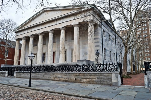 Neoclassical temple-style building with a row of tall stone columns and a decorative iron fence in front, set on a cobblestone street.