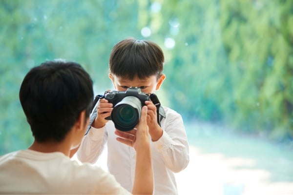 Boy in a white shirt holds a camera up to his face, taking a photo of a person outdoors with a green, sunlit background.