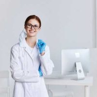 clinical - fundsforNGOs Smiling female scientist in a white lab coat and safety glasses stands in a bright lab, holding a blue glove.