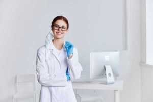Smiling female scientist in a white lab coat and safety glasses stands in a bright lab, holding a blue glove.