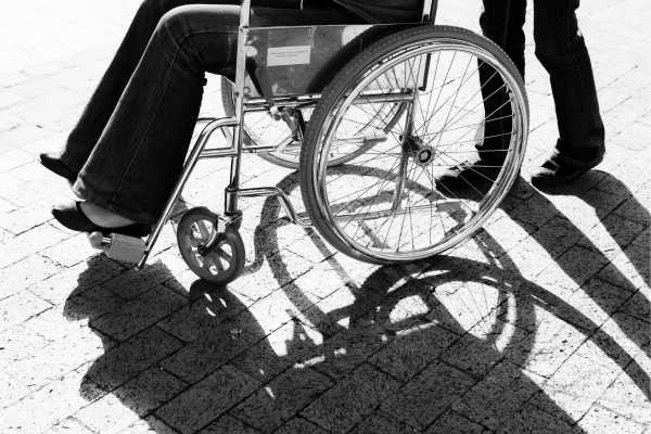 disability 2 - fundsforNGOs Person in a wheelchair being pushed on a sunlit brick sidewalk; strong shadows on the ground.