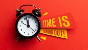Black analog alarm clock on a red surface with bold yellow banners reading 'TIME IS RUNNING OUT!'