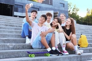 Six teenagers pose for a selfie on outdoor concrete steps, a purple skateboard at their feet and a yellow backpack nearby.