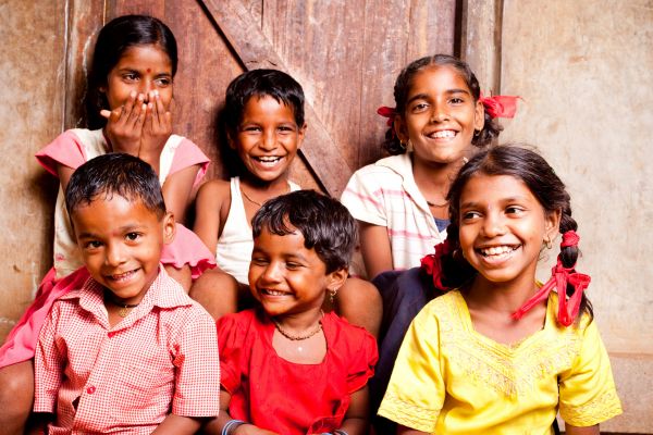 fundsforNGOs - 2026-06-01T132300573 - fundsforNGOs Six smiling children of diverse backgrounds sit closely together in front of a wooden door.