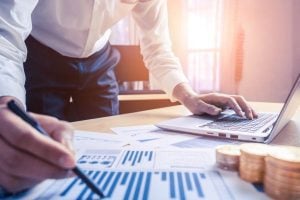 Businessperson examining financial charts on a desk, with a laptop and a stack of coins nearby.