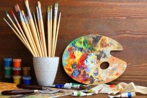 Artist's workspace with paintbrushes in a white cup, a colorful painter's palette, and tubes of paint on a wooden table.