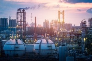 Industrial refinery complex at dusk with smokestacks, storage tanks, and lattice structures bathed in sunset light.
