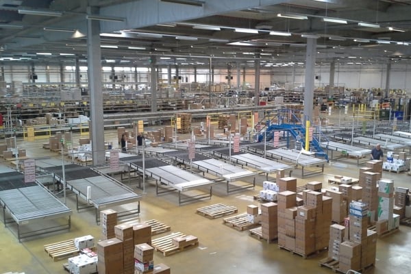 Wide view of a large warehouse with conveyor belts, tall shelves, and stacks of cardboard boxes and pallets.
