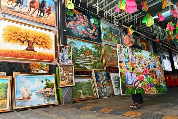 Art vendor carrying a large framed painting among a display of colorful artworks in an open-air market stall.