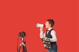 Young boy in a vest speaks into a megaphone beside a film clapboard and a camera on a tripod against a solid red background—film production setup by a kid.