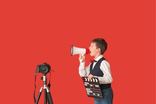 fundsforNGOs - 2026-06-02T125226603 - fundsforNGOs Young boy in a vest speaks into a megaphone beside a film clapboard and a camera on a tripod against a solid red background—film production setup by a kid.