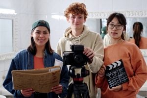 Three students in a studio: left holds a script folder, middle operates a camera on a tripod, right holds a clapperboard.