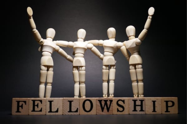 fundsforNGOs - 2026-06-02T130546574 - fundsforNGOs Four wooden mannequins stand side by side with arms linked, atop blocks spelling 'FELLOWSHIP'.