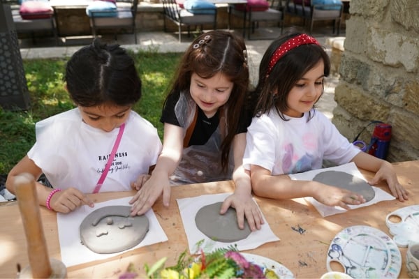 fundsforNGOs - 2026-06-02T132516581 - fundsforNGOs Three girls sit at a sunny outdoor table, shaping gray clay into moon shapes on paper.