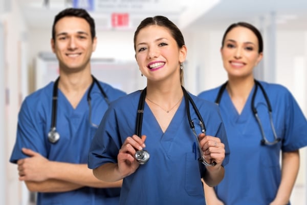Three medical professionals in blue scrubs posing in a bright hospital hallway, with the central nurse smiling and holding a stethoscope around her neck.