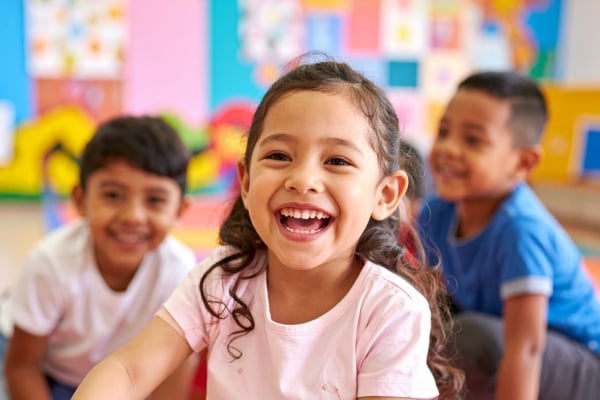 Three smiling children in a colorful classroom, with a girl in a pink shirt laughing center stage and two friends in the background.
