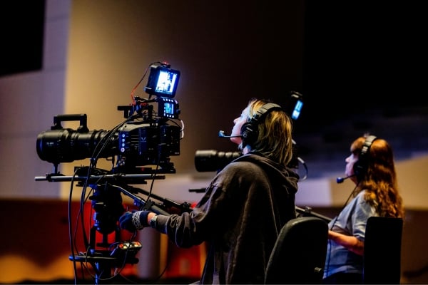 Two video crew members operate a professional camera on a tripod in a dim studio, wearing headsets and monitoring the shot.
