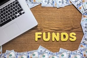 Laptop on a wooden desk with scattered dollar bills and yellow letters spelling FUNDS nearby.